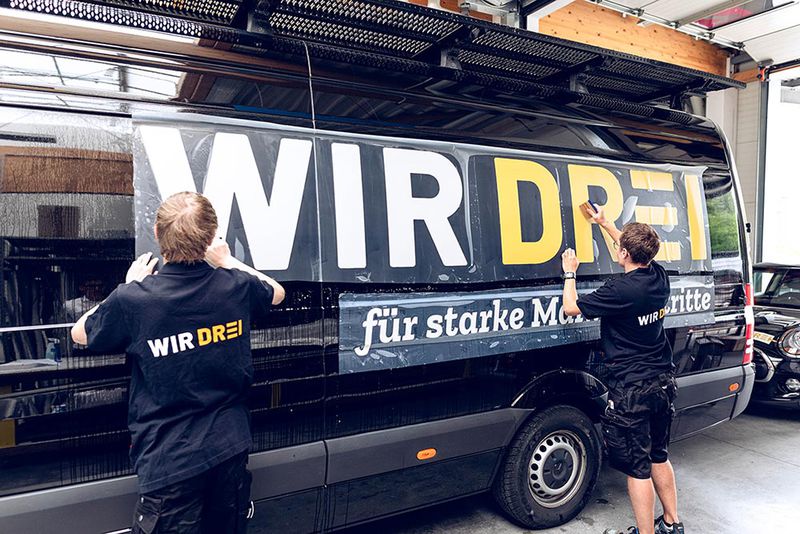 Beklebung eines schwarzen Lieferwagens mit dem WIR DREI Logo durch zwei Mitarbeiter.